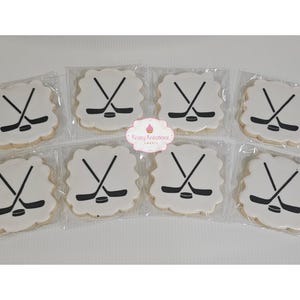 Pode incluir: Oito biscoitos de açúcar branco decorados com desenhos pretos de taco de hóquei e disco. Os biscoitos são embalados individualmente em sacos de plástico transparentes.
