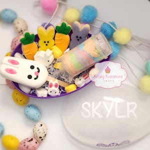 May include: An Easter-themed gift set featuring a purple basket filled with bunny and carrot-shaped treats, a push-pop candy, and decorative eggs. A white egg-shaped container with the name "SKYLR" is also present, along with a garland of colorful pom-poms.