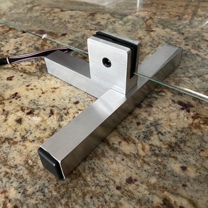 May include: A silver metal stand with a black rubber foot, designed to hold a glass panel. The stand has a T-shaped base and a vertical piece with a clamp to secure the glass.