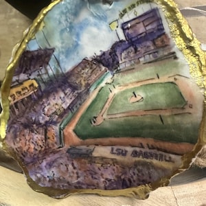 May include: Hand-painted oyster shell ornament featuring a baseball field scene with the text "LSU Baseball." The shell has a gold-painted edge and a white ribbon for hanging. The artwork includes a stadium and a green field.