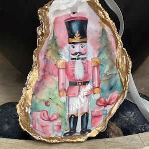 Pode incluir: Enfeite de Natal pintado à mão em concha de ostra, apresentando um soldado quebra-nozes em uniforme vermelho e chapéu preto. A concha tem uma borda dourada e uma fita branca para pendurar. O fundo inclui árvores de Natal e presentes embrulhados.