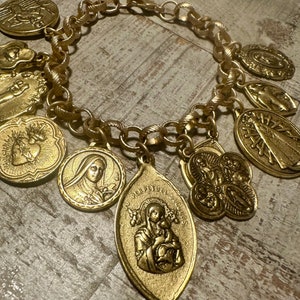 May include: A gold chain bracelet with multiple charms, including religious medallions and a heart. The charms are all gold and have intricate designs.