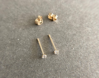 Könnte beinhalten: Ein Paar goldene Ohrstecker mit kleinen, klaren Edelsteinen. Die Ohrringe liegen auf einer grauen Oberfläche.