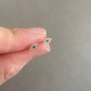 May include: A pair of gold-colored stud earrings, each shaped like an eye. The earrings feature a blue center stone and are surrounded by small, clear stones. The earrings are held between fingers.