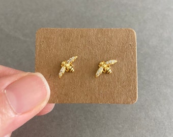 May include: A pair of gold bee stud earrings with clear crystal accents. The earrings are displayed on a brown card.