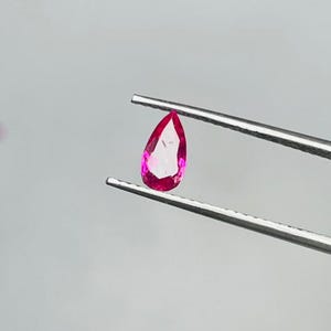 May include: A pear-shaped, vibrant pink gemstone is held by a pair of silver tweezers against a light gray background. The gemstone has a faceted cut, reflecting light. A small, similar gemstone is visible in the background.