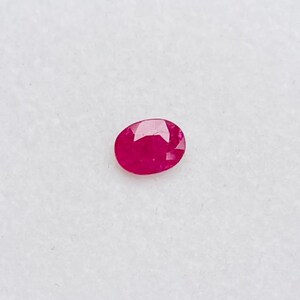 May include: A vibrant, oval-cut ruby gemstone. The ruby is a deep, rich red color and is set against a white background. The gemstone is displayed in a small, clear plastic container.