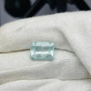 Peut inclure: Une pierre précieuse rectangulaire bleu clair avec une qualité translucide subtile. La gemme est présentée sur un fond de tissu blanc, soulignant sa clarté et sa couleur. La taille de la pierre est précise, mettant en valeur ses facettes.