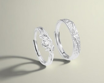 May include: Two silver rings. One ring features a floral design with a central gemstone and delicate leaves. The other ring has a textured, bark-like pattern. Both rings are set against a neutral background, showcasing their intricate details.