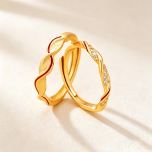Puede incluir: Dos anillos dorados con un diseño trenzado en forma de onda. Un anillo presenta una fina incrustación de esmalte rojo a lo largo de la curva, mientras que el otro está adornado con una serie de pequeñas piedras redondas y claras.