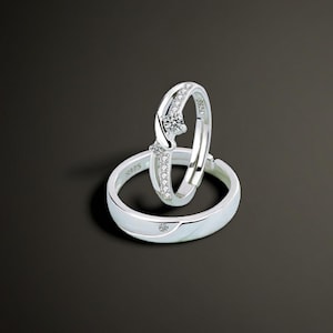 Può includere: Due anelli d'argento dal design minimalista. Un anello presenta un diamante centrale e pietre più piccole lungo la fascia. L'altro anello ha un singolo diamante. Entrambi gli anelli sono su uno sfondo scuro.