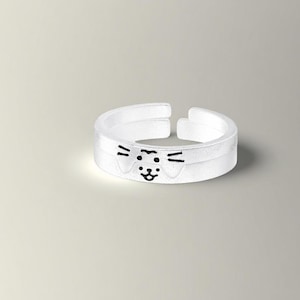 Op de afbeelding: Een heldere, verstelbare ring met een kattengezichtontwerp. De ring heeft een witte band met een zwarte kattengezichtsillustratie. Het kattengezicht heeft twee zwarte lijnen voor snorharen, twee zwarte lijnen voor ogen en een zwarte neus en mond.