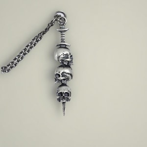Può includere: Una collana con pendente argentato con tre ciondoli a forma di teschio impilati. I teschi presentano tratti del viso dettagliati e sono collegati da una barra verticale. La collana include una catena e un elemento superiore decorativo.