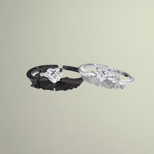 Puede incluir: Dos anillos ajustables, uno negro y otro plateado, cada uno con una piedra preciosa en forma de corazón. Los anillos están diseñados con un motivo de pluma. El anillo plateado tiene un acabado pulido, mientras que el anillo negro tiene un acabado mate. Ambos anillos están sobre un fondo neutro.