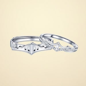 Puede incluir: Un par de anillos de tono plateado. Un anillo presenta un diseño geométrico que recuerda a la visera de un casco de caballero con aberturas verticales. El segundo anillo tiene una banda ondulada tipo corona con una pequeña gema clara en forma de pera.