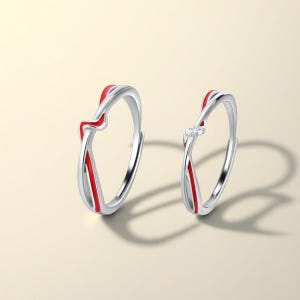 Peut inclure: Deux bagues argentées au design torsadé et fin, ornées d'une incrustation en émail rouge vif. L'une des bagues présente une ligne rouge lisse et continue, tandis que l'autre est agrémentée de trois petites pierres claires et brillantes.