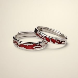 May include: Two silver rings with a red enamel design. The rings have a smooth, polished finish and a band of red enamel that wraps around the exterior. The rings are likely adjustable, with one slightly open. The rings are set against a neutral background.