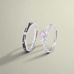May include: Two silver rings. One ring features a black band with star cutouts and a small clear stone. The other ring has a pink star-shaped stone, a crescent moon, and small clear stones.