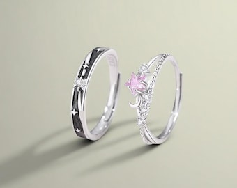 May include: Two silver rings. One ring features a black band with star cutouts and a small clear stone. The other ring has a pink star-shaped stone, a crescent moon, and small clear stones.