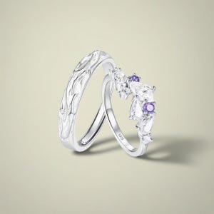 Könnte beinhalten: Zwei silberne Ringe. Ein Ring hat ein strukturiertes Design, der andere eine gebogene Reihe weißer und violetter Edelsteine. Die Ringe sind vor einem hellen Hintergrund platziert.