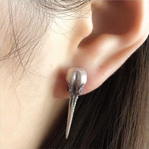 Puede incluir: Un pendiente plateado con forma de cráneo de pájaro. El pendiente es de tipo botón, con una parte superior detallada en forma de cráneo y un pico largo y puntiagudo. El pendiente se lleva en una oreja, centrándose en la joya.