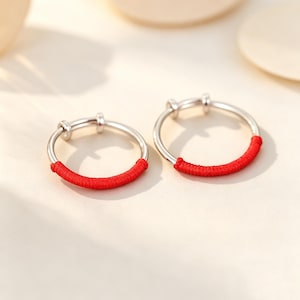 Peut inclure: Deux bagues de couleur argentée avec un design circulaire. Chaque bague est partiellement enveloppée d'un fil rouge vif, couvrant environ un tiers de la circonférence. Les anneaux métalliques ont une finition lisse et polie et une structure ouverte.