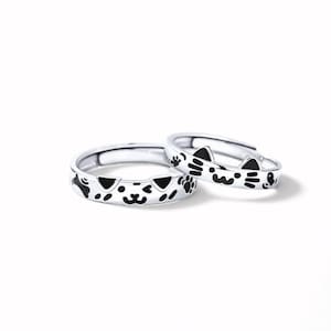 May include: Two silver-toned adjustable rings featuring black enamel details. One ring displays a cat face with whiskers and ears, while the other ring shows a dog face with ears, a bone shape, and paw prints. Both rings have an open band design.