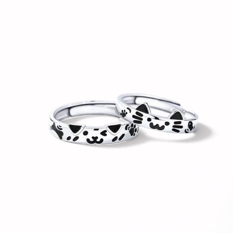 May include: Two silver-toned adjustable rings featuring black enamel details. One ring displays a cat face with whiskers and ears, while the other ring shows a dog face with ears, a bone shape, and paw prints. Both rings have an open band design.
