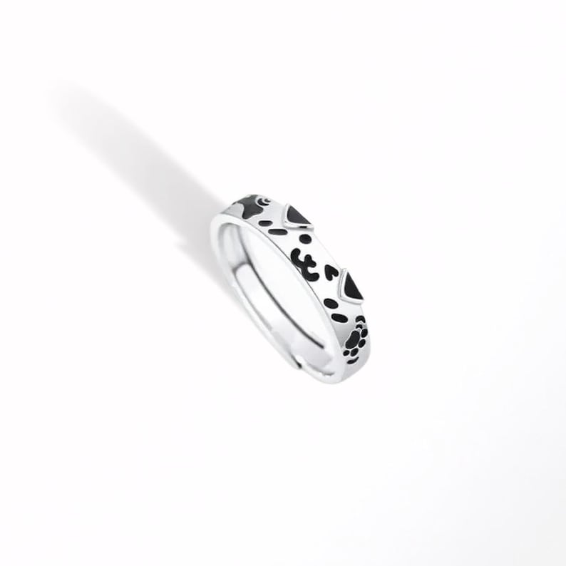 May include: A silver-toned ring featuring a repeating pattern of black enamel paw prints and small heart shapes around the outer band. The ring has an open-ended design, allowing for adjustment, and displays a polished metallic finish throughout.