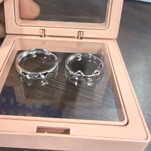 May include: Two silver-toned metal rings with open bands, one featuring a repeating heart cutout pattern and the other a cat-ear design, displayed inside a square, light pink plastic case with a clear hinged lid and a dark interior base.
