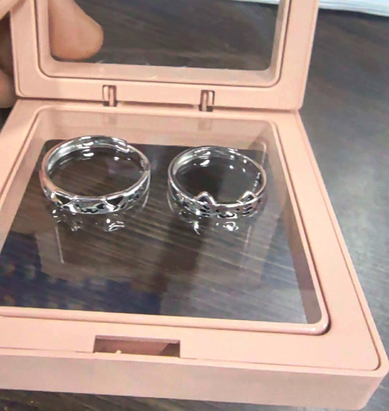May include: Two silver-toned metal rings with open bands, one featuring a repeating heart cutout pattern and the other a cat-ear design, displayed inside a square, light pink plastic case with a clear hinged lid and a dark interior base.