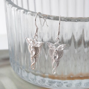 May include: A pair of silver earrings with a clear crystal design. The earrings are shaped like a stylized tree with three leaves and a single crystal at the center of each leaf.