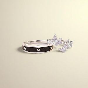 May include: A collection of silver rings featuring butterfly motifs. One ring has a black band with butterfly cutouts. The other rings have butterfly-shaped details with small, clear gemstones. The rings are set against a light beige backdrop.