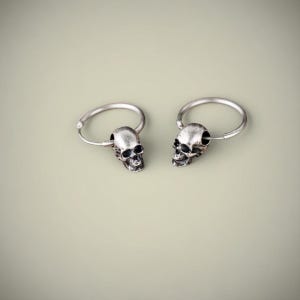 May include: A pair of silver-toned hoop earrings, each featuring a small, detailed skull charm suspended from the circular wire band. The metal has a darkened, oxidised finish in the recessed areas to highlight the facial features of the skulls.