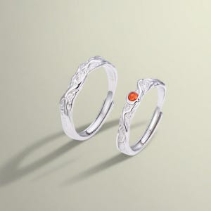 Può includere: Due anelli d'argento con un design organico e strutturato. Un anello presenta una piccola gemma rotonda arancione. Gli anelli sono posti su uno sfondo neutro, evidenziando i loro dettagli intricati e l'elegante semplicità.