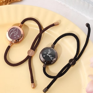 Pode incluir: Duas pulseiras ajustáveis com um cordão preto e um amuleto redondo. O amuleto à esquerda é de ouro rosa com um desenho branco de um pássaro com estrelas. O amuleto à direita é preto com um desenho branco de uma cordilheira com estrelas.