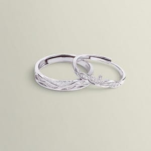 Puede incluir: Dos anillos de plata. Uno presenta un diseño tejido, mientras que el otro tiene un patrón de hojas con pequeños acentos brillantes. Los anillos se colocan sobre un fondo suave y neutro, resaltando su elegante diseño.