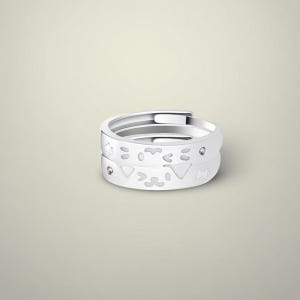 Puede incluir: Dos anillos blancos ajustables apilados uno encima del otro. El anillo inferior presenta formas recortadas, incluyendo una cara de gato y otros diseños geométricos. Cada anillo tiene una pequeña gema transparente. Los anillos son brillantes.