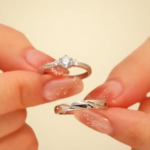Puede incluir: Se muestran dos anillos de plata. Un anillo presenta una piedra preciosa central transparente con piedras más pequeñas a cada lado. El otro anillo tiene un diseño retorcido con un acabado pulido. Ambos anillos son sostenidos por una persona.