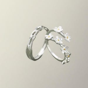 Puede incluir: Dos anillos de plata, uno con un delicado diseño floral y el otro con un patrón más intrincado en forma de rama. El anillo floral presenta pequeñas flores blancas con centros amarillos.