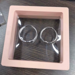 May include: Two silver-toned hoop earrings are displayed inside a square, light pink floating frame case. The earrings are circular with a polished metallic finish and a hinged closure mechanism, positioned side-by-side within the clear display.