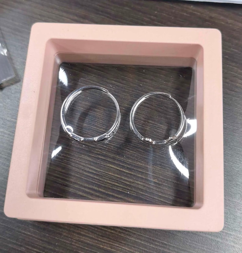 May include: Two silver-toned hoop earrings are displayed inside a square, light pink floating frame case. The earrings are circular with a polished metallic finish and a hinged closure mechanism, positioned side-by-side within the clear display.