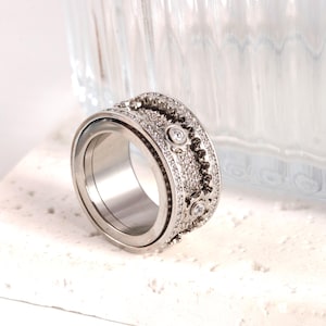Puede incluir: Un anillo giratorio de plata con un diseño decorativo que presenta pequeñas piedras transparentes y engranajes. El anillo está sobre una superficie blanca.