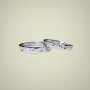 May include: Two silver-toned rings featuring a textured surface with engraved four-pointed star motifs. The rings are positioned with one slightly overlapping the other, showcasing a metallic finish and a consistent band width throughout the design.