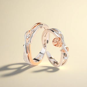 Puede incluir: Dos anillos de oro rosa y plata. Uno presenta una rosa y hojas, el otro un diseño de vid. Los anillos están sobre un fondo amarillo suave. Es probable que los anillos sean ajustables.