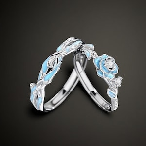 Può includere: Un anello in argento con un design a vite scolpito, accenti in smalto azzurro e una piccola gemma trasparente incastonata in un dettaglio a forma di rosa. L'anello ha una finitura metallica lucida e una struttura organica complessa.