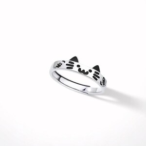 May include: A silver-toned metal ring featuring a cat-themed design with black enamel detailing. The band displays two pointed cat ears at the top, along with a stylized face, whiskers, and a small paw print graphic etched into the metal surface.