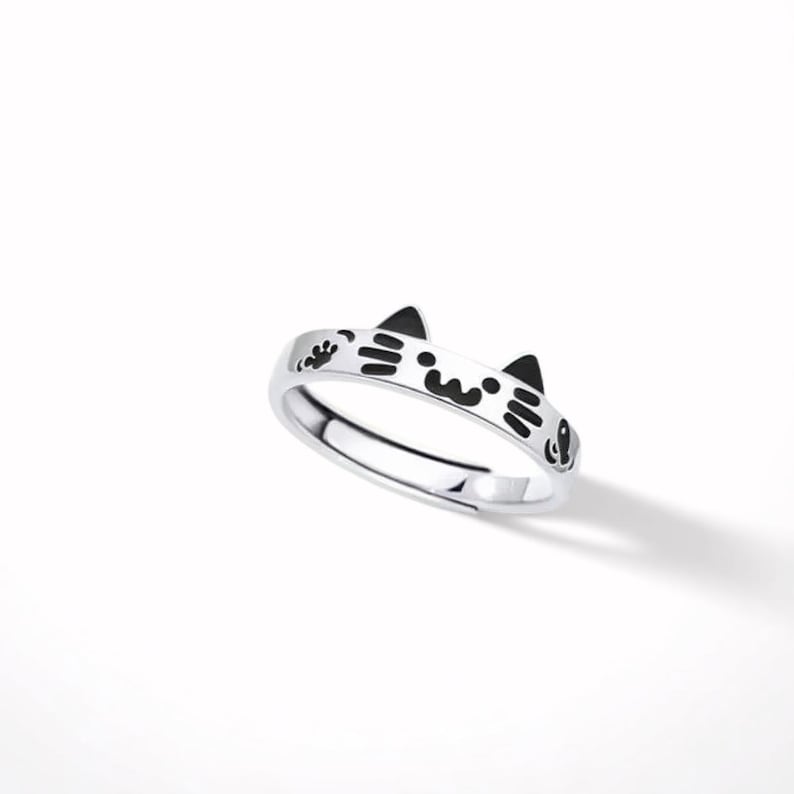 May include: A silver-toned metal ring featuring a cat-themed design with black enamel detailing. The band displays two pointed cat ears at the top, along with a stylized face, whiskers, and a small paw print graphic etched into the metal surface.