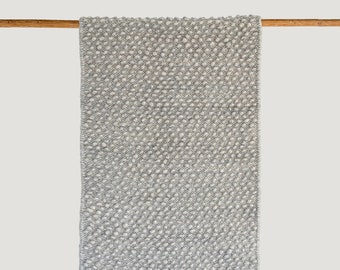 May include: A rectangular, gray and white textured rug hanging from a wooden rod. The rug has a woven pattern with a mix of light and dark gray threads, creating a subtle, speckled appearance. The rug is displayed against a plain, off-white background.