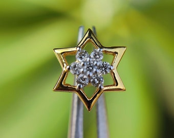 May include: A gold star-shaped pendant with a cluster of small diamonds in the center. The pendant is held by two black-tipped tweezers.